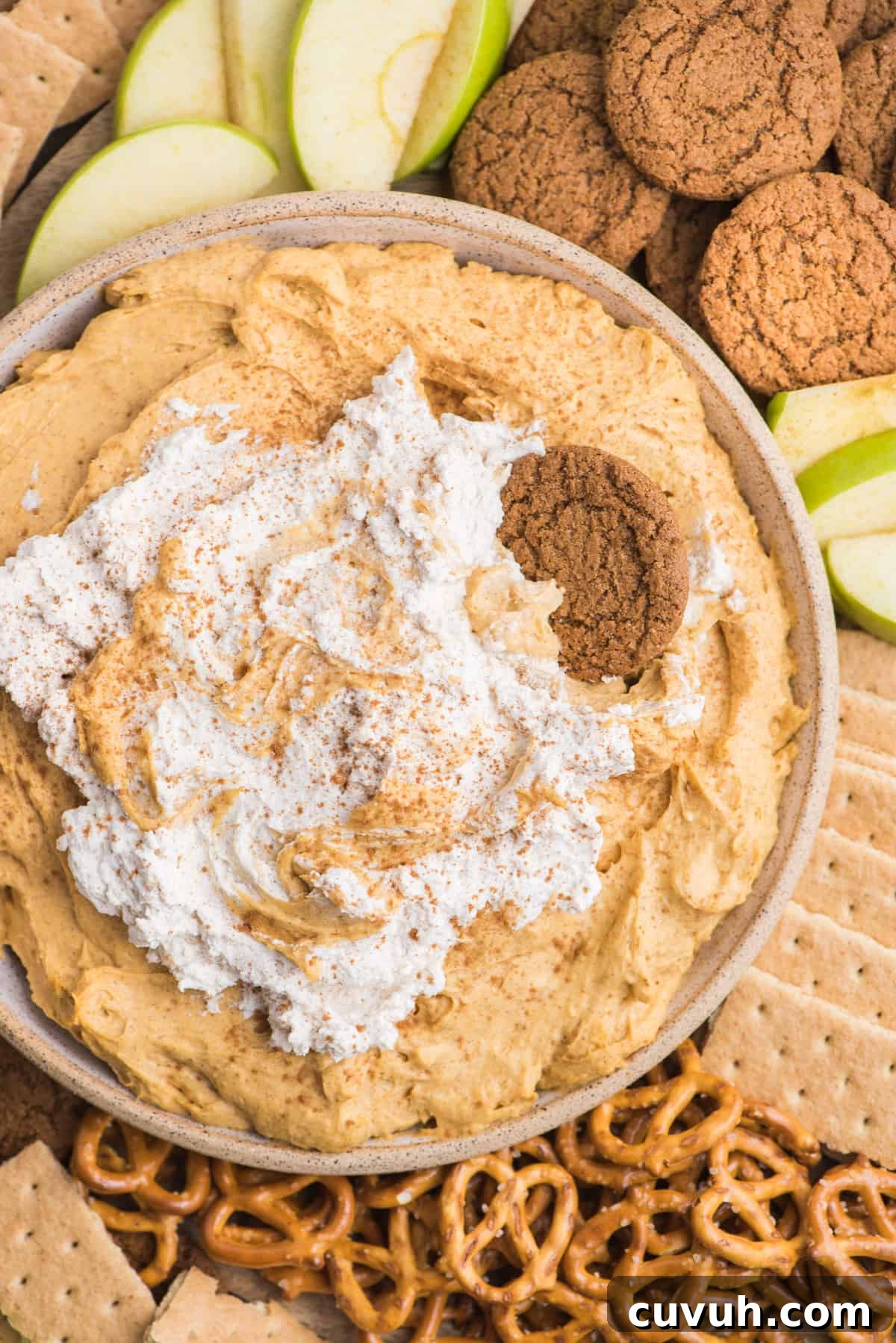 pumpkin dip with whipped cream topping in a bowl surrounded by piles of pretzels, gingersnaps, graham crackers and green apple slices