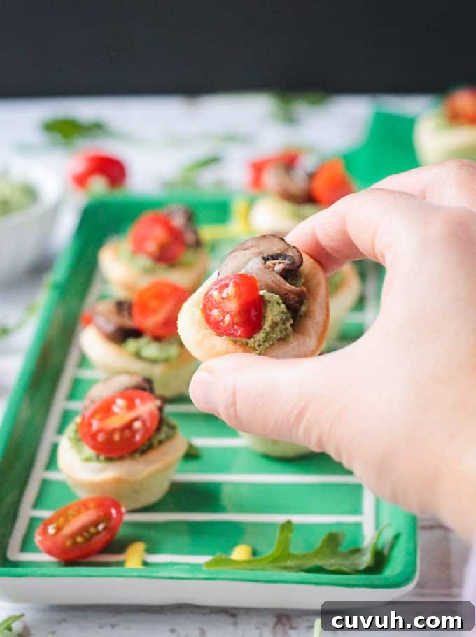 Vegan Pizza Bites - easy pizza dough topped with dairy free pesto, mushrooms and cherry tomatoes. Big bold flavors in a little bite - perfect for your next party or get together! Especially great for game day! Hand holding one vegan pizza bite. Tray full of more pizza bites in the background.