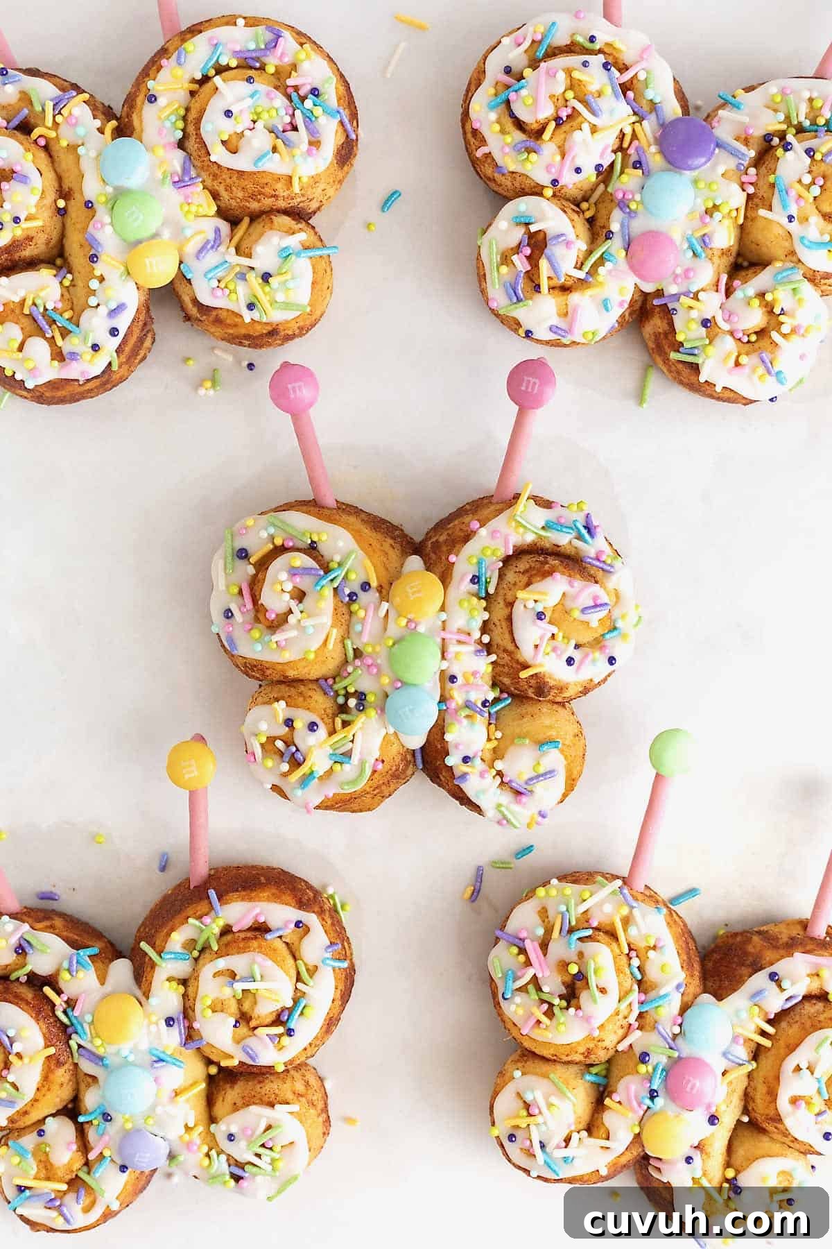 Five butterfly shaped cinnamon rolls decorated with icing, pastel candies and candy antennae on a whit surface.