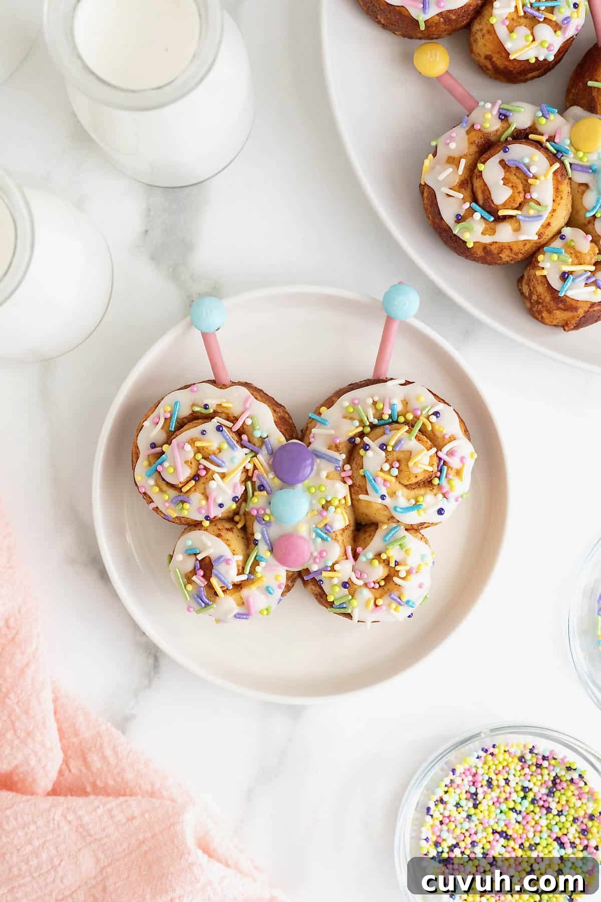 A butterfly-shaped cinnamon roll decorated with white icing and pastel M&Ms and sprinkles on a white plate.