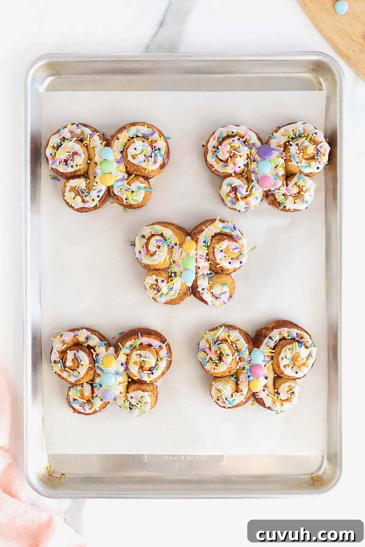 Five butterfly shaped cinnamon rolls decorated with white icing and pastel candies on a parchment lined aluminum baking sheet.