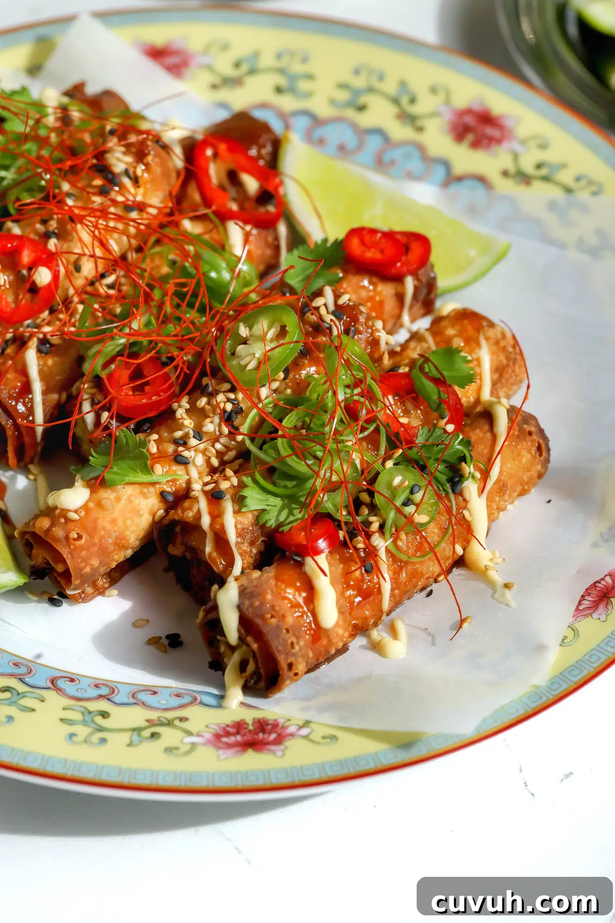 Crab Rangoon Taquitos with Sweet Chili Honey Glaze 2 Close-up side view of crispy crab Rangoon taquitos piled on a decorative plate, drizzled with sweet chili honey and lemon mayo, and garnished with sesame seeds, cilantro, sliced chiles, and red chili threads.