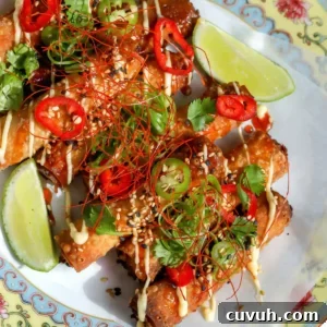 Crab Rangoon Taquitos with Sweet Chili Honey Glaze 12 Top-down view of golden fried crab Rangoon taquitos on a parchment-lined plate, topped with lemon mayo, sweet chili honey, sliced jalapenos, red chiles, fresh cilantro, and sesame seeds.