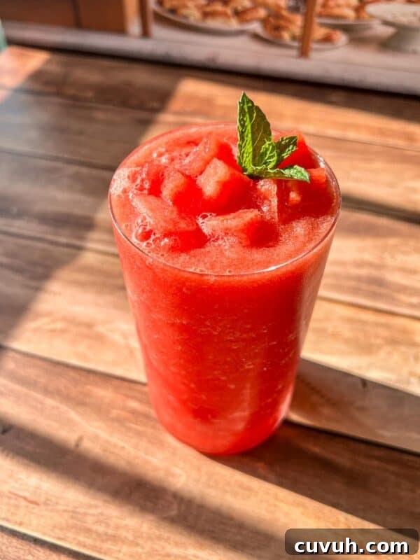 A glass of bright red watermelon smoothie topped with small watermelon cubes and a sprig of fresh mint.