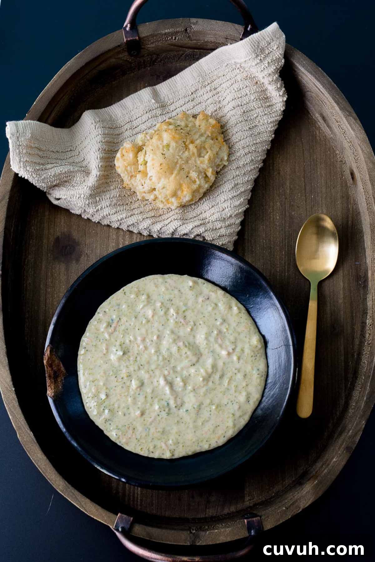 Soup served in black bowl on wooden platter with gold spoon and biscuit overhead shot