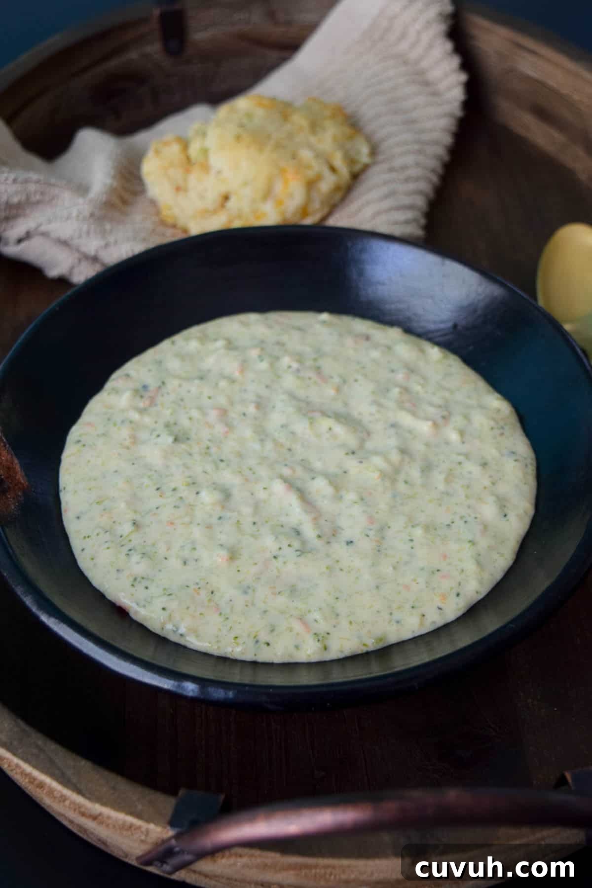 The Best Broccoli Cheddar Soup in black bowl with biscuit behind bowl on brown napkin close up view
