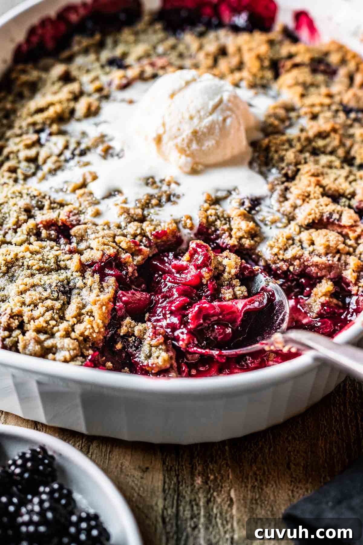 Apple and Blackberry Crumble Recipe for Perfect Autumn Dessert 9 A spoon full of fruit crumble in a casserole dish with a scoop of vanilla ice cream in the middle.