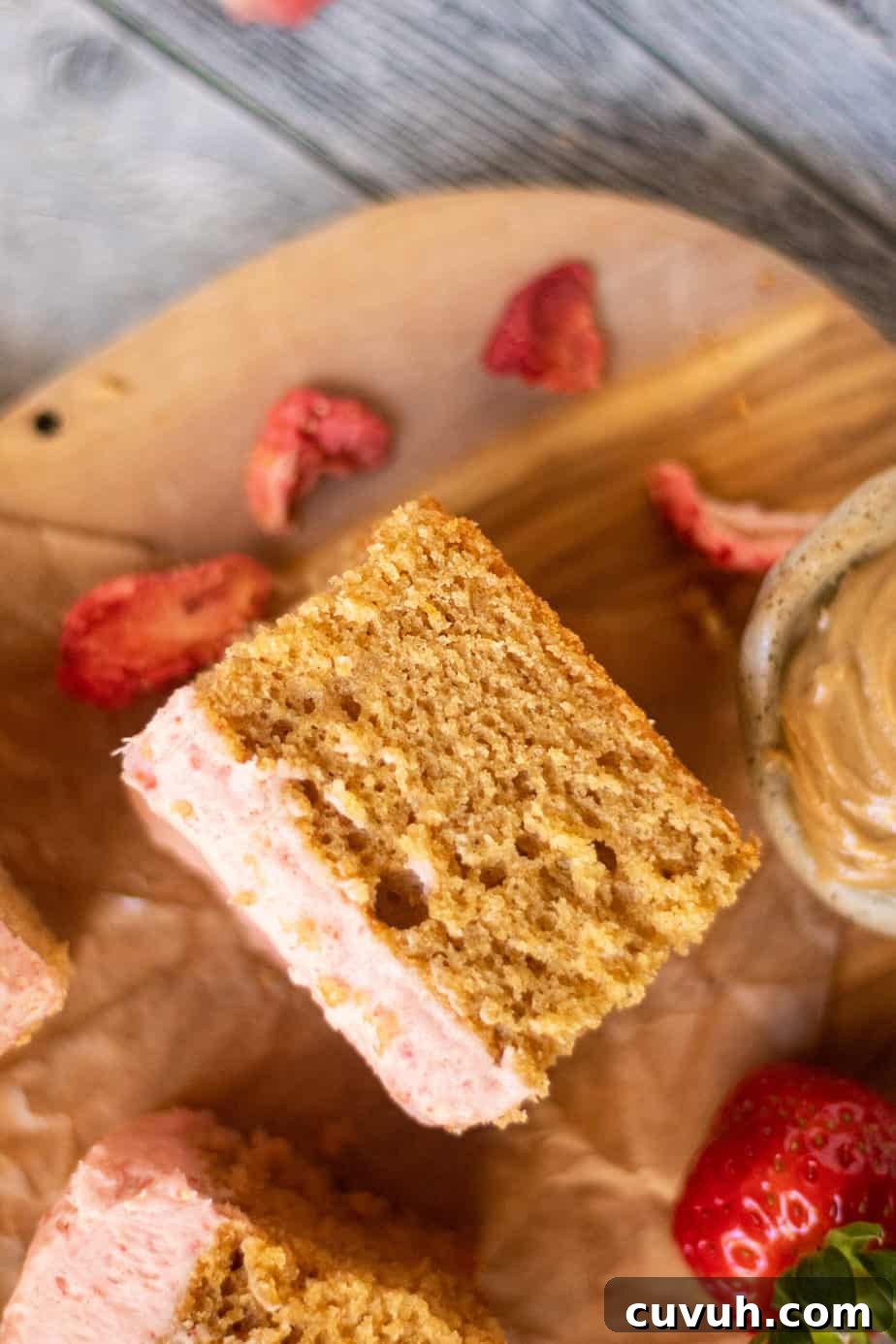 Peanut Butter Layer Cake with Strawberry Buttercream Frosting 10 close up overhead shot of sliced peanut butter cake with strawberry frosting on a wooden cutting board. sliced strawberries scattered in perimeter of shot.