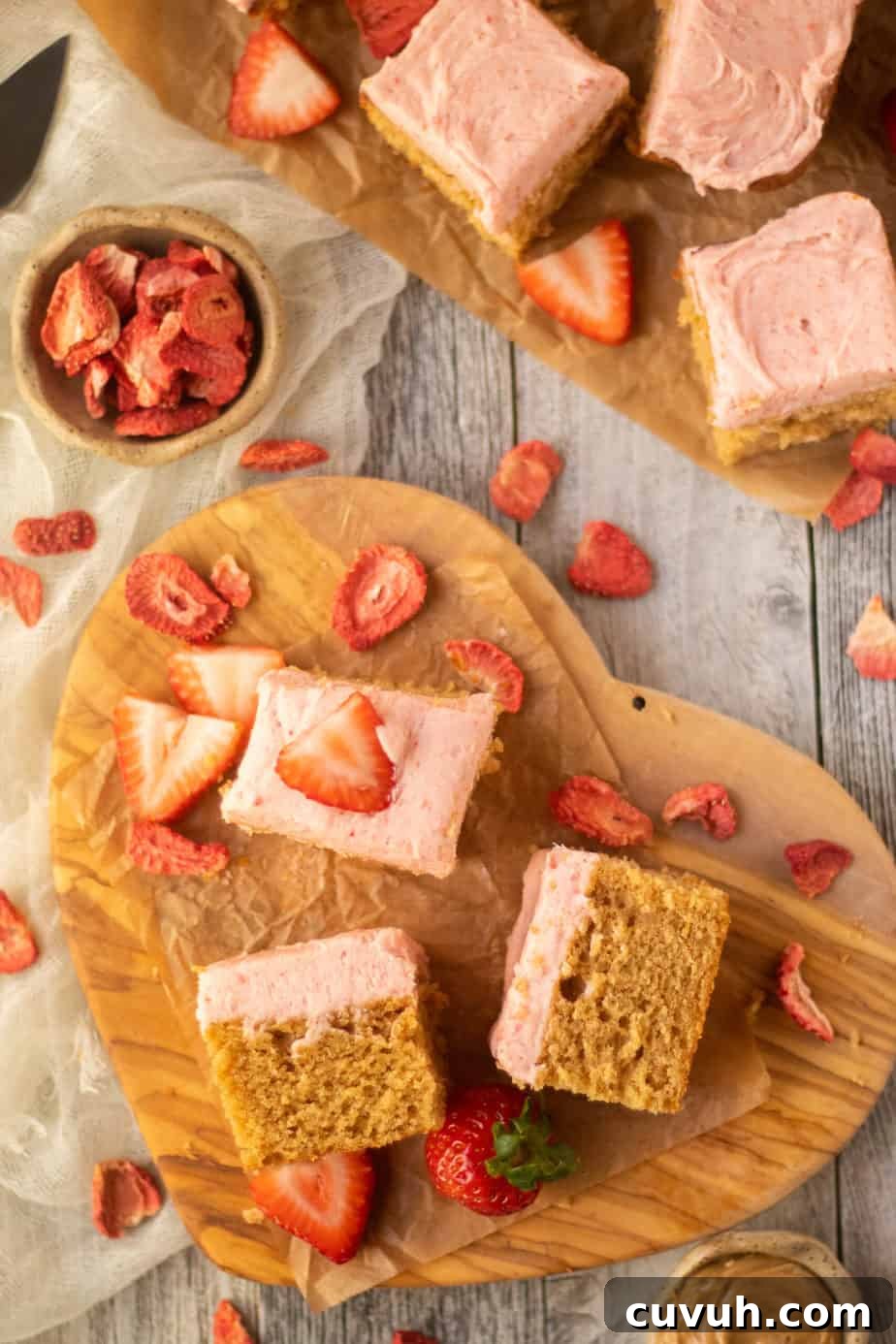 Peanut Butter Layer Cake with Strawberry Buttercream Frosting 3 overhead shot of sliced peanut butter cake with strawberry frosting on a wooden cutting board. sliced strawberries scattered in perimeter of shot.