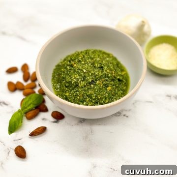 Almond Pesto Sauce Recipe: Fresh Basil & Parmesan Blend 3 Bright green almond pesto in a white bowl on a white marble counter garnished with fresh basil leaves and an almond