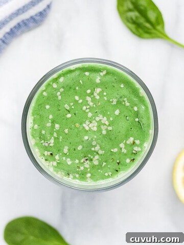 A green smoothie in a clear glass, garnished with white hemp seeds. There is half a lemon, 3 spinach leaves, and a blue and white striped dish cloth in the background.