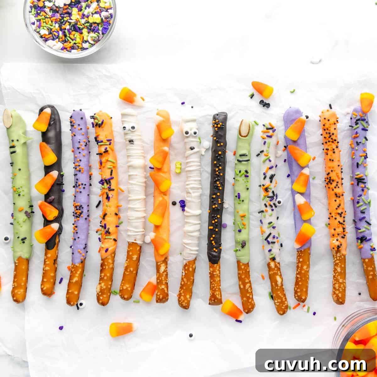 A variety of pretzel sticks covered in different colored coatings and decorated with candy eyes, sprinkles, and candy corn, arranged on a white surface.