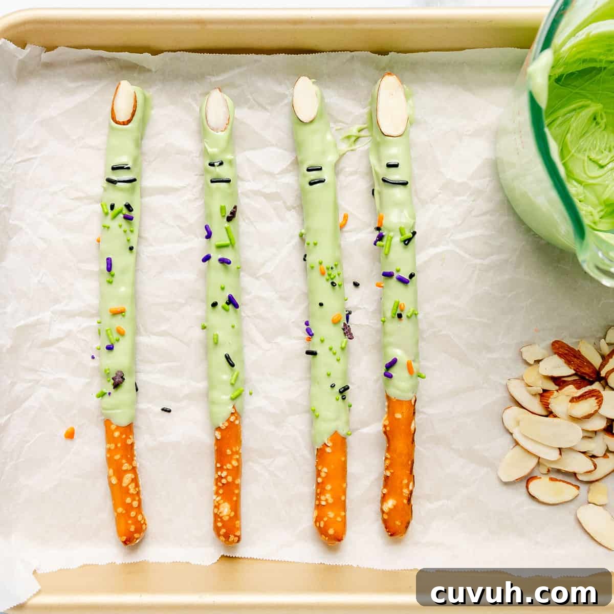 Four pretzel rods dipped in green chocolate, decorated with sprinkles and almond slivers, arranged on parchment paper next to a bowl of green chocolate and a pile of almond slivers.