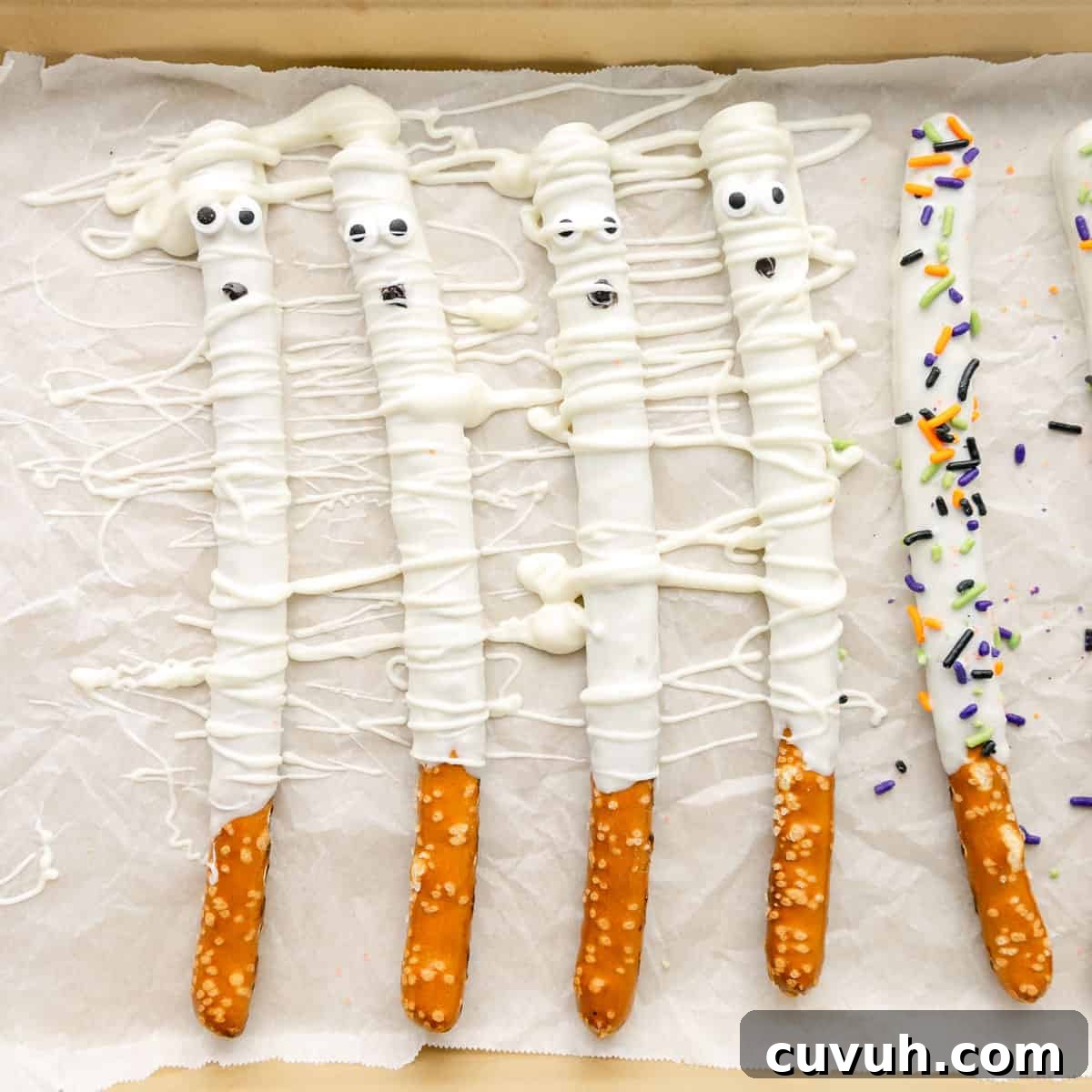 Pretzel rods coated in white chocolate and decorated to resemble mummies, with candy eyes and sprinkles, arranged on parchment paper.