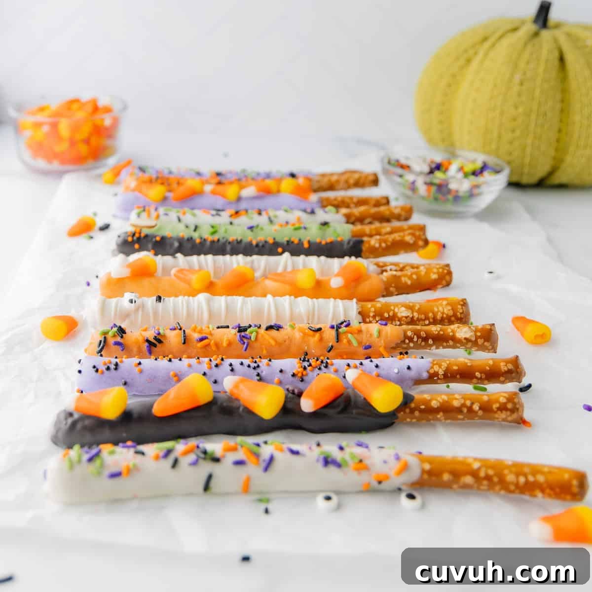 Pretzel rods coated in various colored icing and decorated with candy corn and sprinkles are arranged on a white surface, with a knitted pumpkin and bowls of more candy and sprinkles in the background.