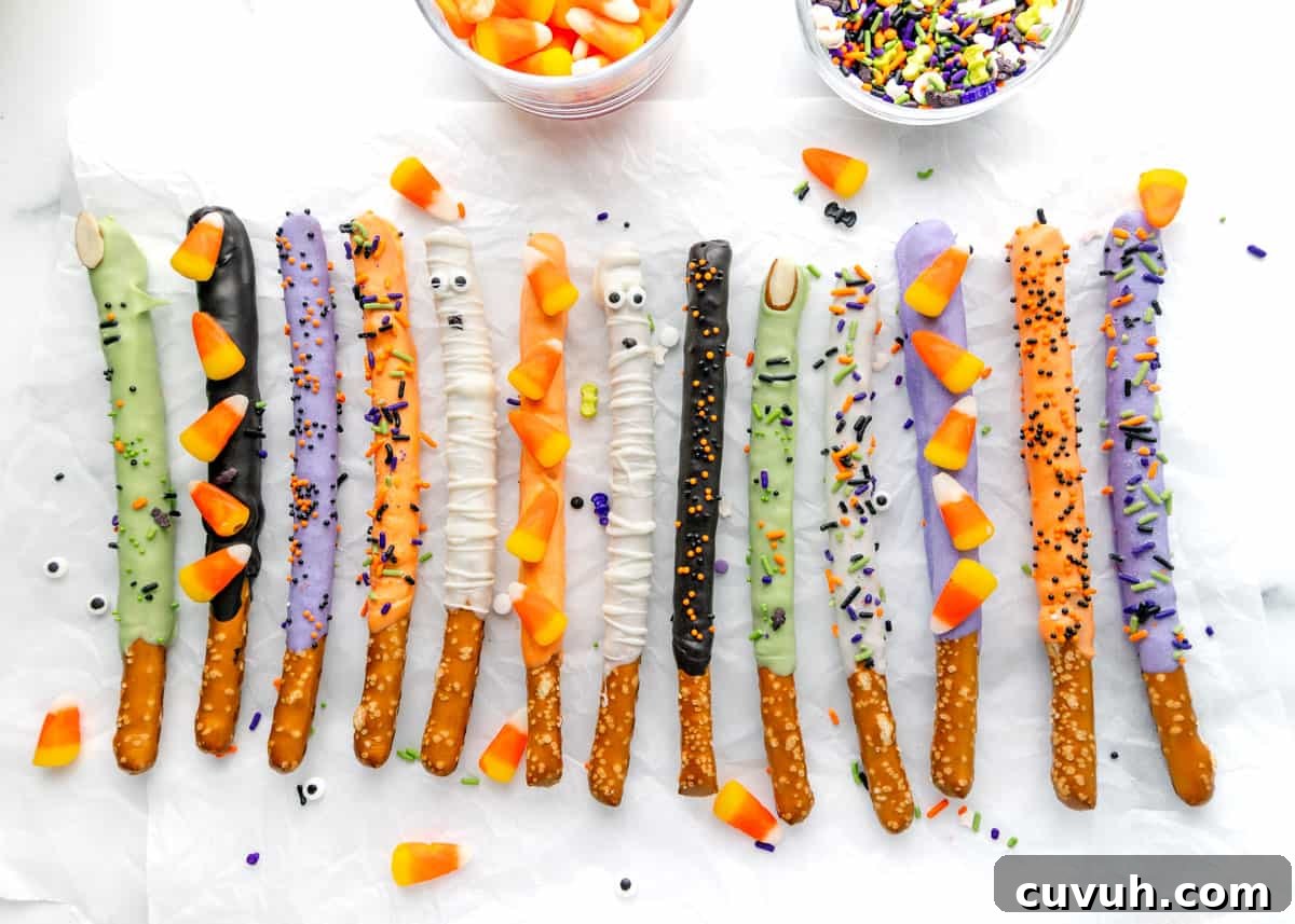 A variety of pretzel sticks dipped in different colored icing and decorated with sprinkles, candy eyes, and candy corn arranged in a row on a white surface.