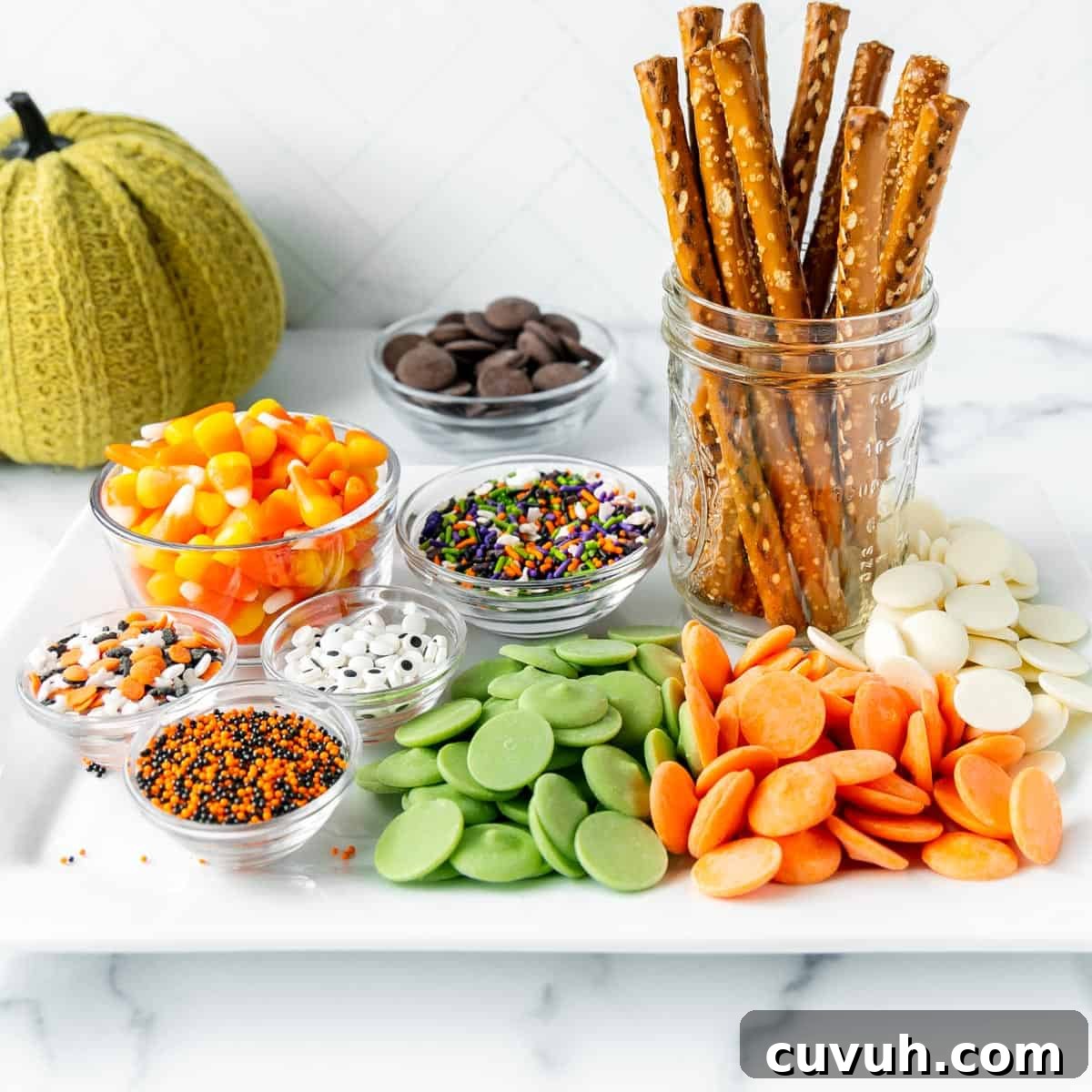 A tray with assorted toppings: pretzel sticks in a jar, candy corn, chocolate chips, multicolored sprinkles, white and green candy melts, and green, black, orange sprinkles. A knit pumpkin decor.
