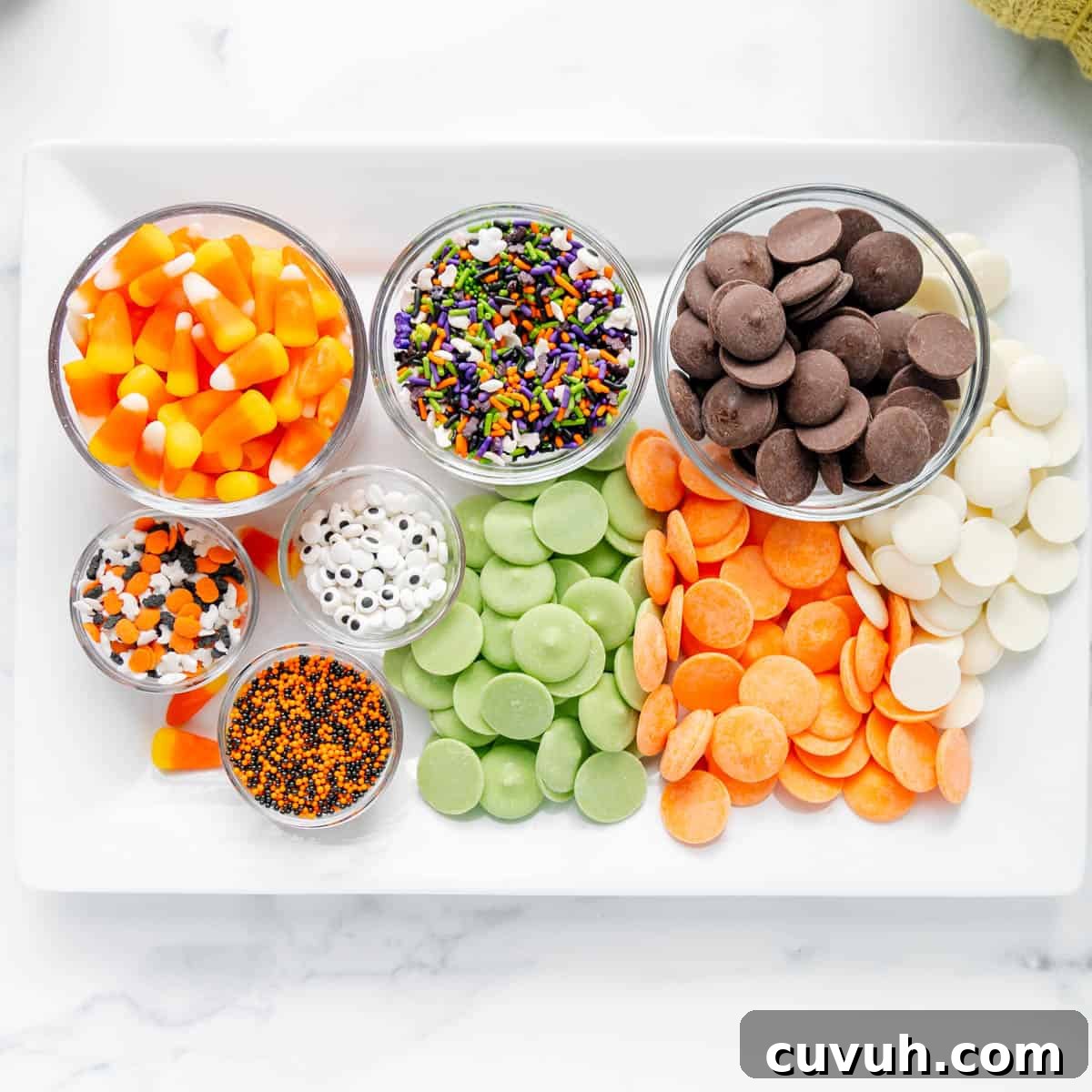 A white tray displaying bowls of candy corn, assorted sprinkles, candy eyeballs, and various colored chocolate melts in green, orange, white, and dark brown.