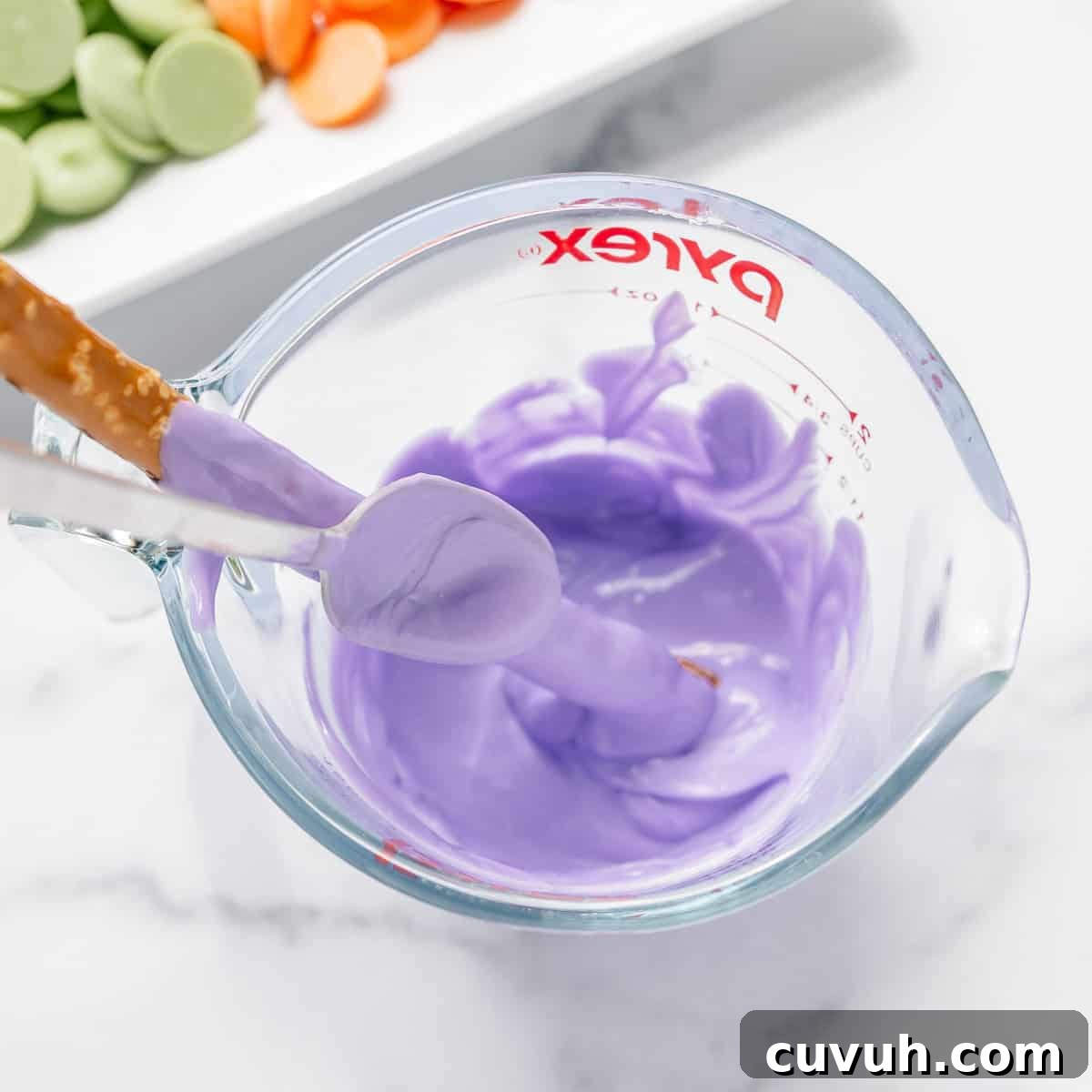 A glass measuring cup filled with melted purple candy coating, with a spatula and dipped pretzel stick inside. In the background, green and orange candy melts are on a white tray.