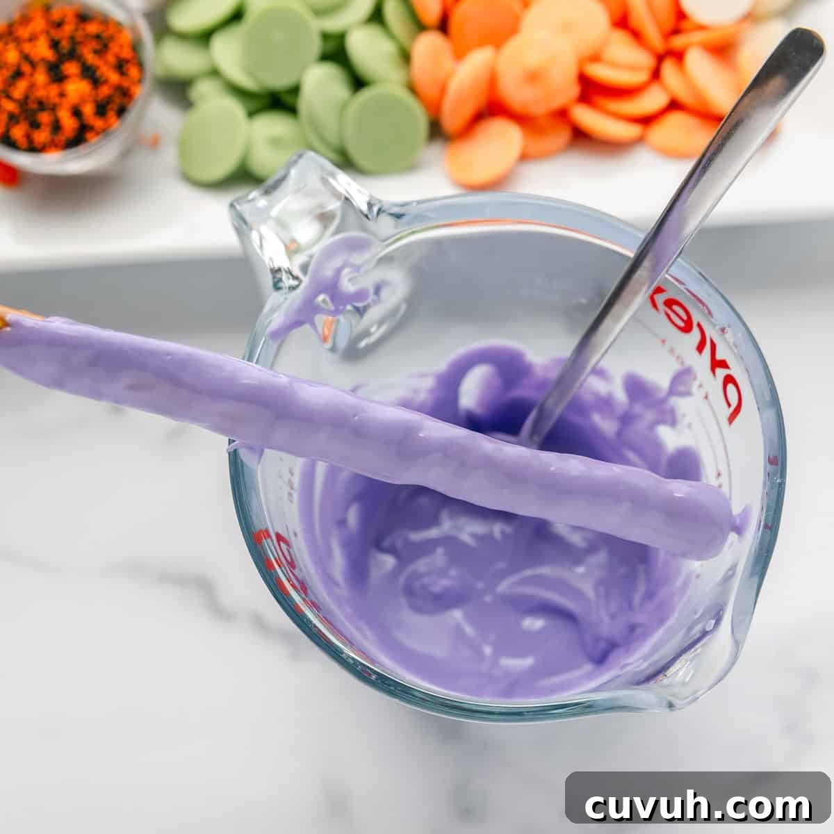 A glass measuring cup filled with melted purple candy coating, a pretzel rod being dipped in the coating, and trays of various colored candy melts in the background.