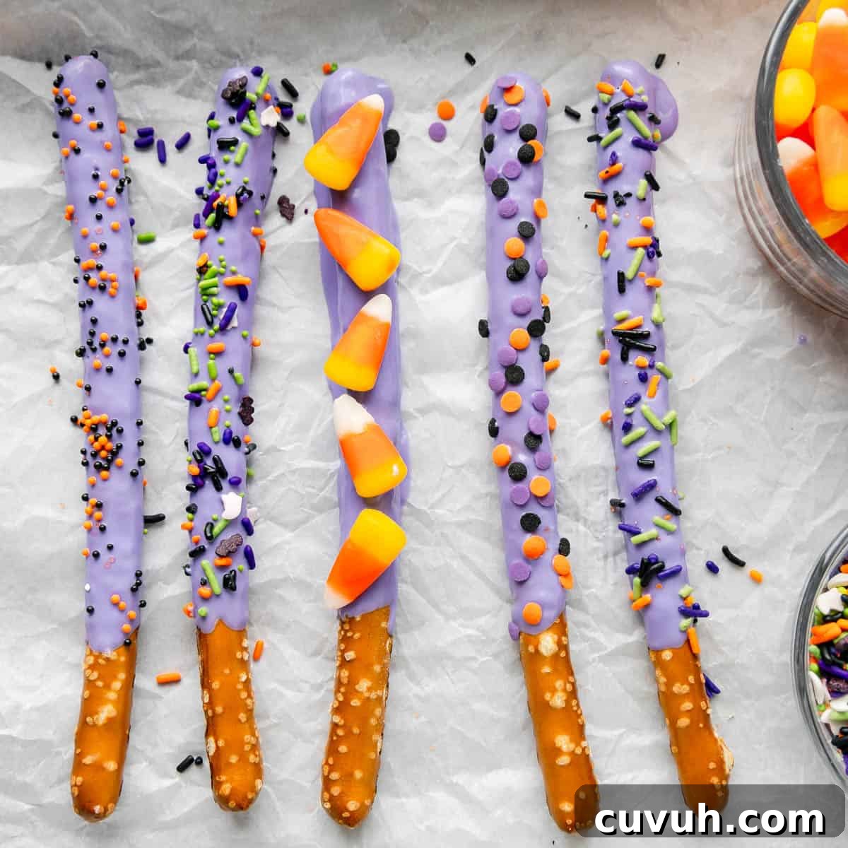 Five pretzel sticks dipped in purple candy coating and decorated with colorful sprinkles and candy corn, arranged in a row on parchment paper.