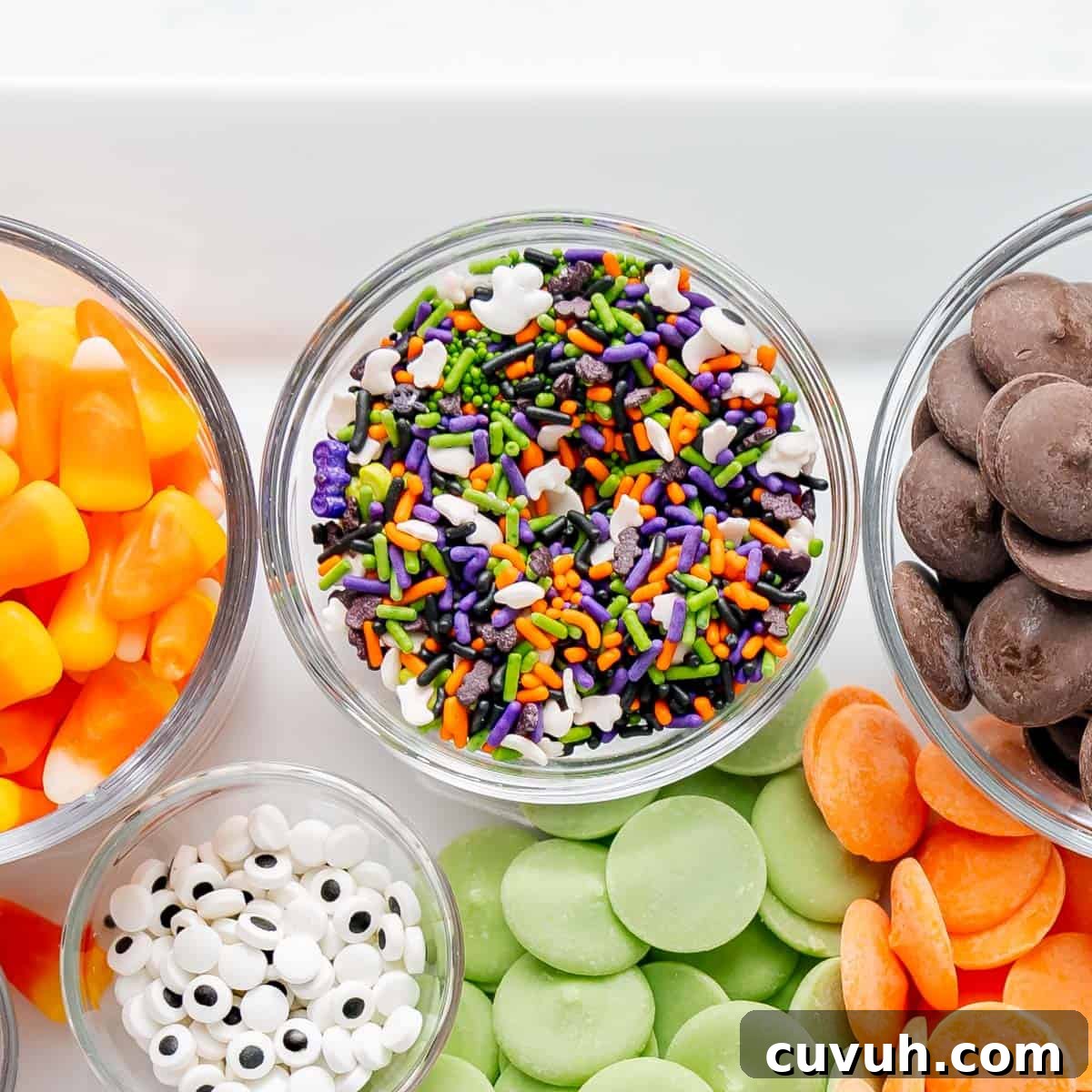 Assorted Halloween-themed treats, including candy corn, colorful sprinkles, candy eyes, chocolate wafers, and orange and green candy melts displayed in bowls.