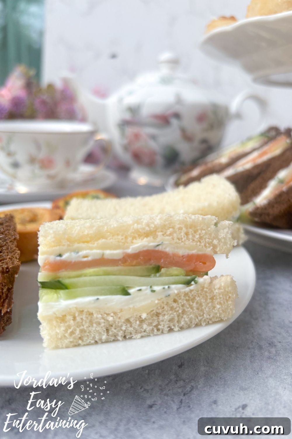 Smoked Salmon Tea Sandwiches with Cream Cheese, Cucumber & Dill 11 Smoked salmon tea sandwiches with cream cheese, cucumber, and dill on a plate with mini quiches and crostini, served alongside floral china tea cups and a matching teapot for an afternoon tea party.