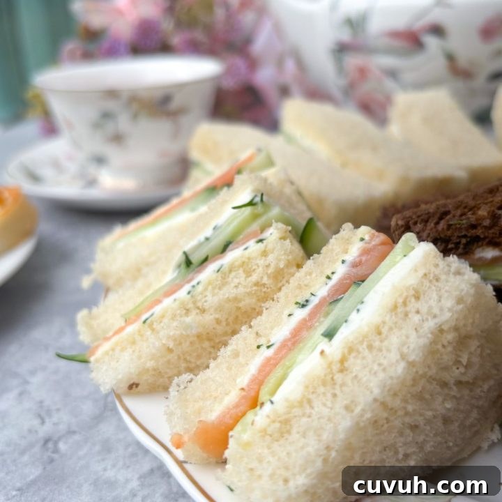 Smoked salmon tea sandwiches with cream cheese, cucumber, and dill on a plate with mini quiches and crostini, served alongside floral china tea cups and a matching teapot for an afternoon tea party.