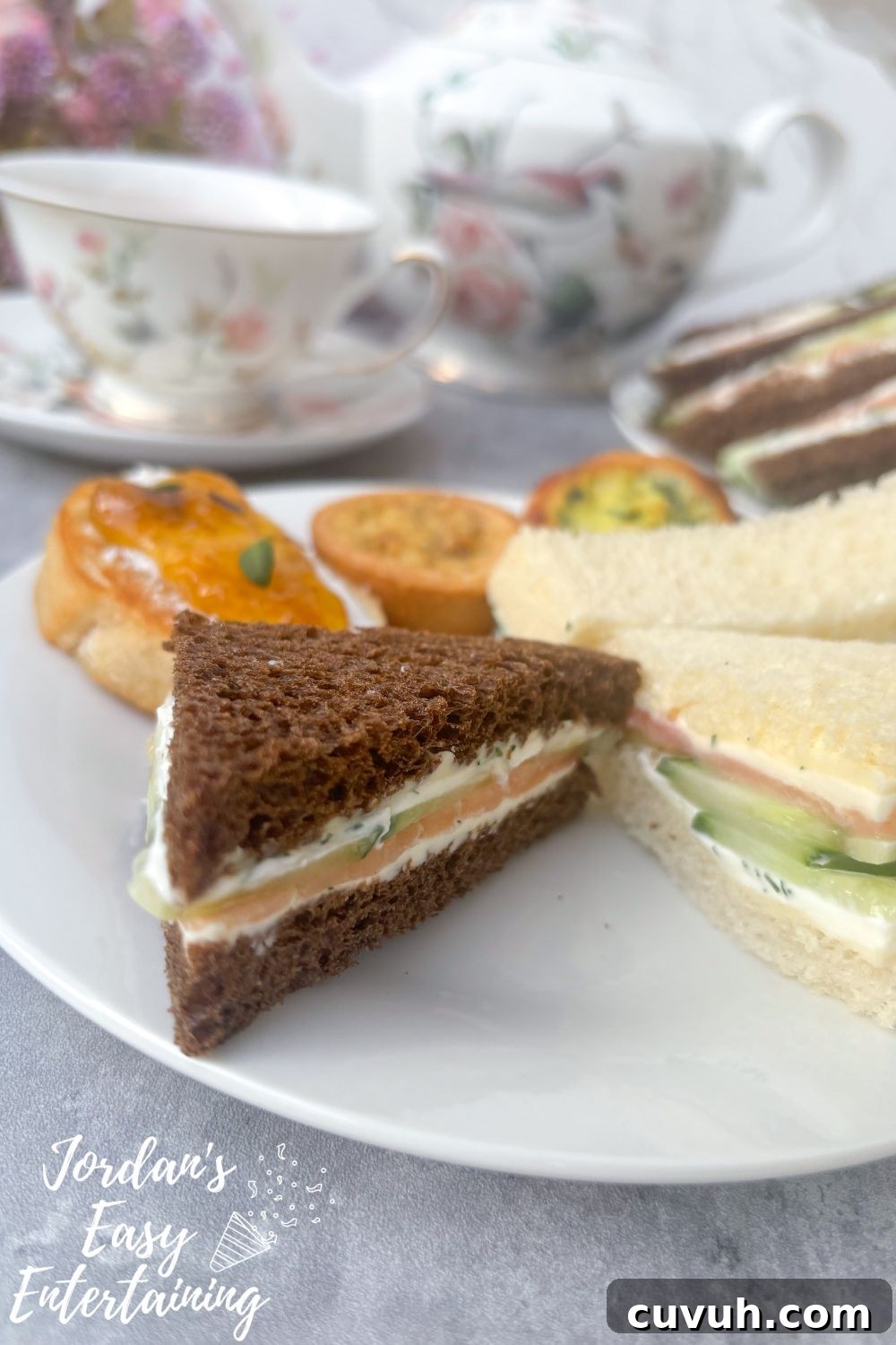 Smoked Salmon Tea Sandwiches with Cream Cheese, Cucumber & Dill 4 Close-up of smoked salmon tea sandwich on pumpernickel bread with cream cheese, cucumber, and dill, served with mini quiches and floral teacups for an elegant afternoon tea.