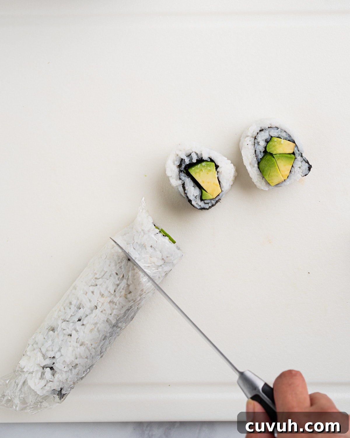 Avocado roll wrapped in plastic wrap and knife cutting into bite size pieces.
