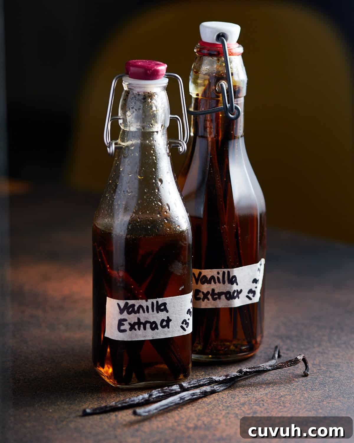 Photo of two small bottles of homemade vanilla extract on a dark worktop with two vanilla pods lying in front.