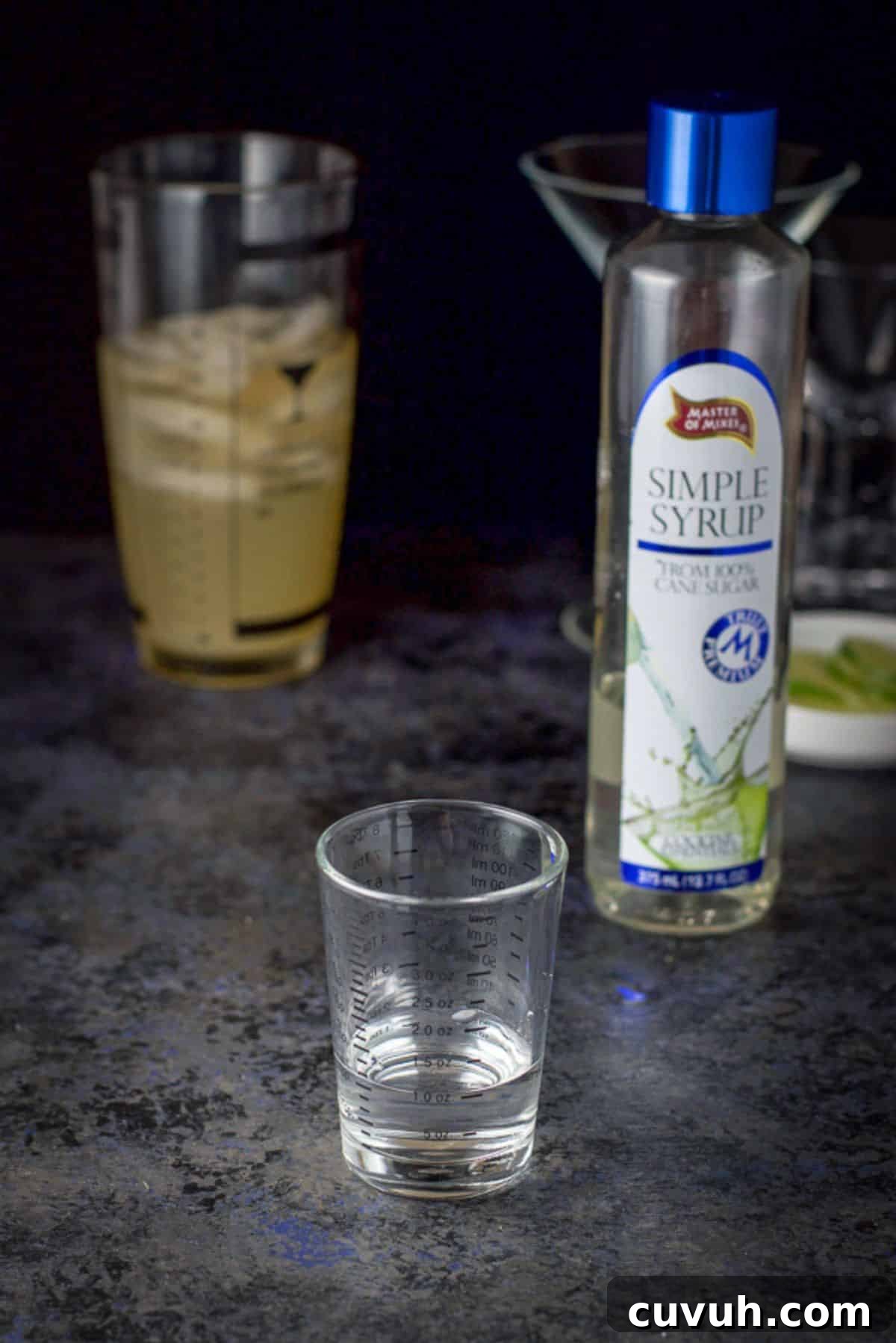 Simple syrup measured out with the bottle in the background and the shaker and glasses in the back as well